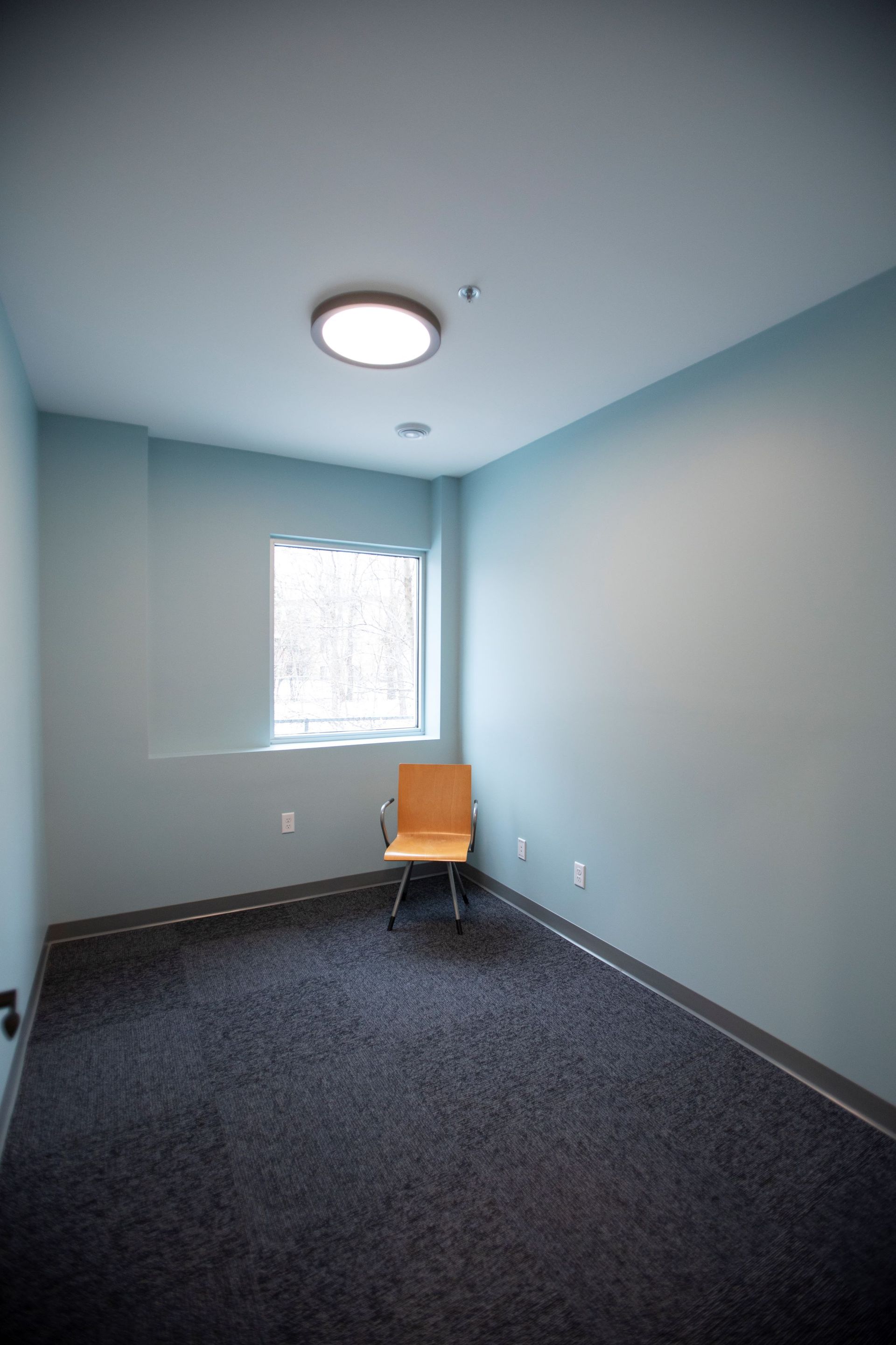 Small room with light blue walls, gray carpet, a window, a chair, and a ceiling light.