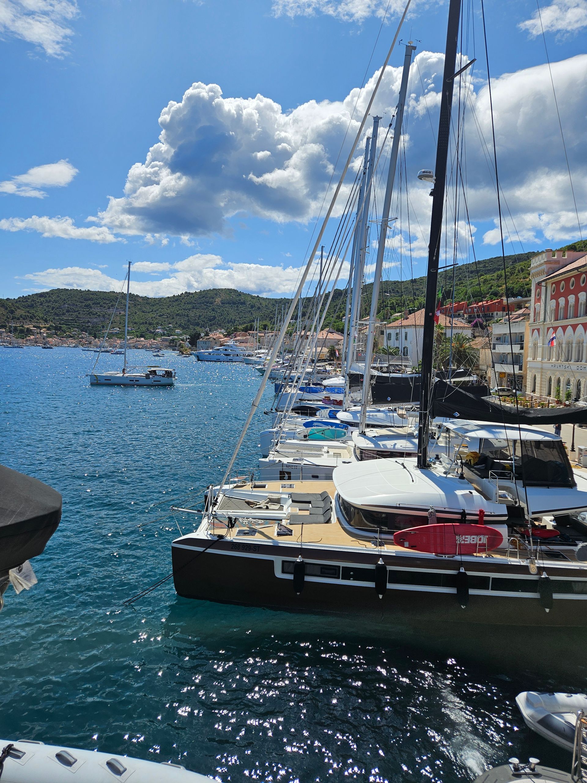 Sailboats docked in a harbor on a sunny day with buildings lining the shore and puffy white clouds in the blue sky.