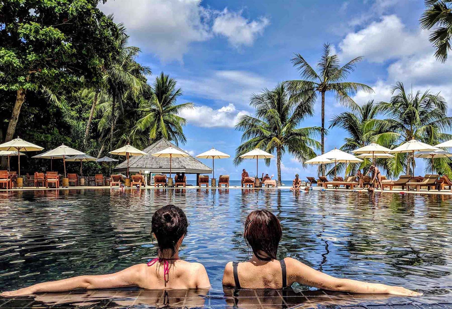 Two women are sitting in a swimming pool with palm trees in the background.