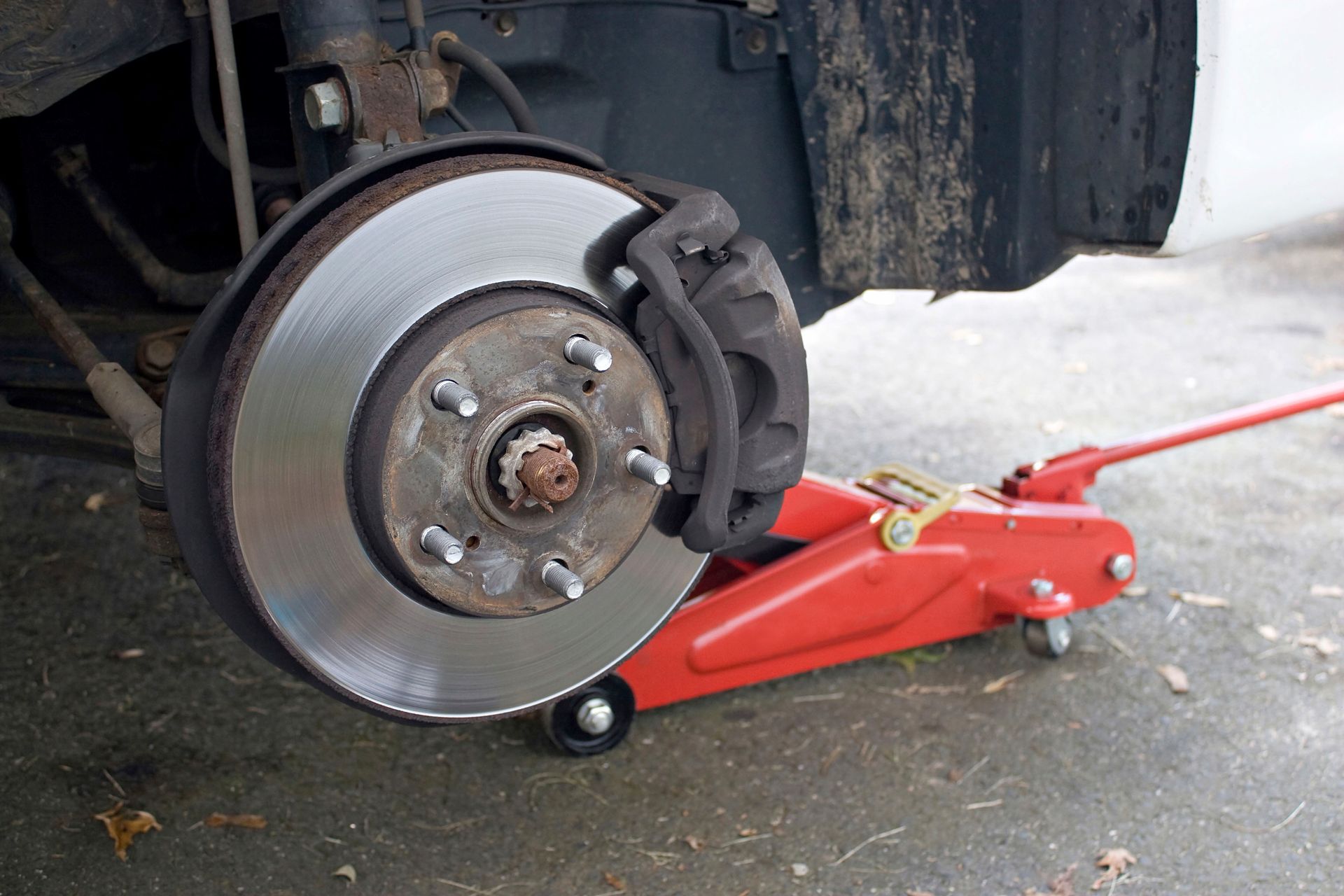 Car brake rotor, caliper, and lug nuts with tire removed, raised by a red jack on concrete.