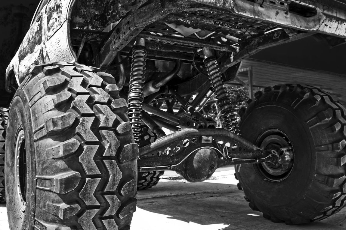 Black and white close-up of a monster truck's suspension and massive tire. The tires are heavily treaded.