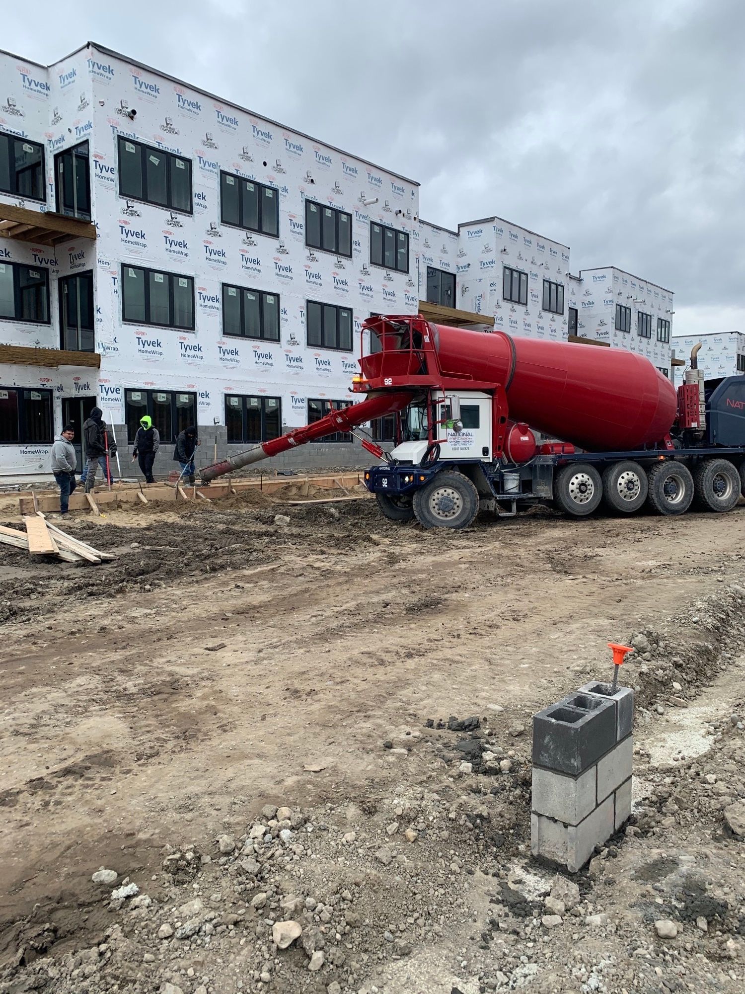 A concrete mixer truck is driving down a dirt road in front of a building under construction.