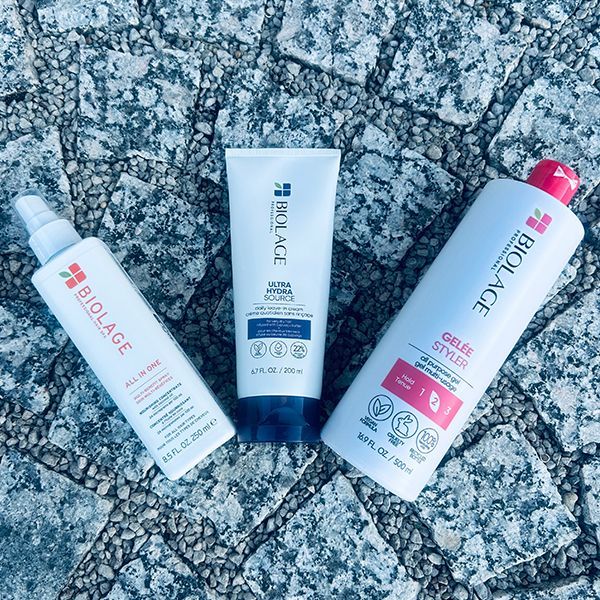 Three Biolage hair products on a stone surface: a spray, a tube, and a large gel bottle.