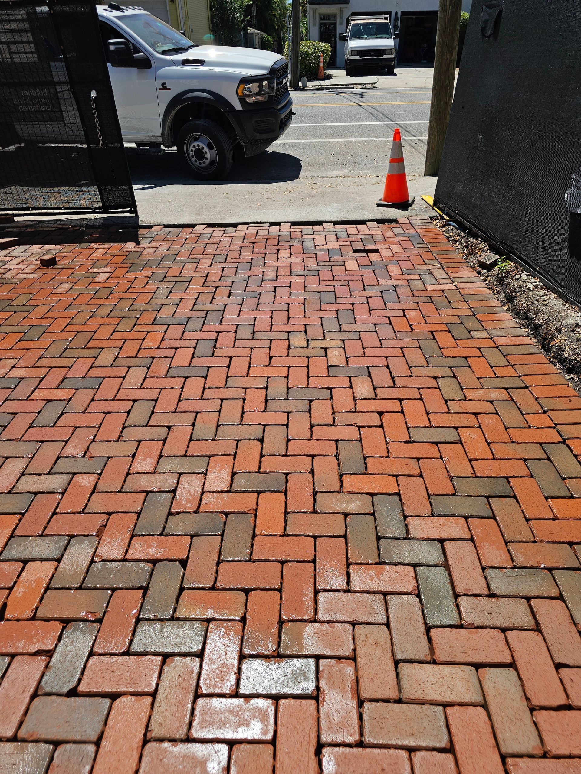 Red brick walkway beside a wall, leading to a parking lot with a traffic cone and parked vehicles.