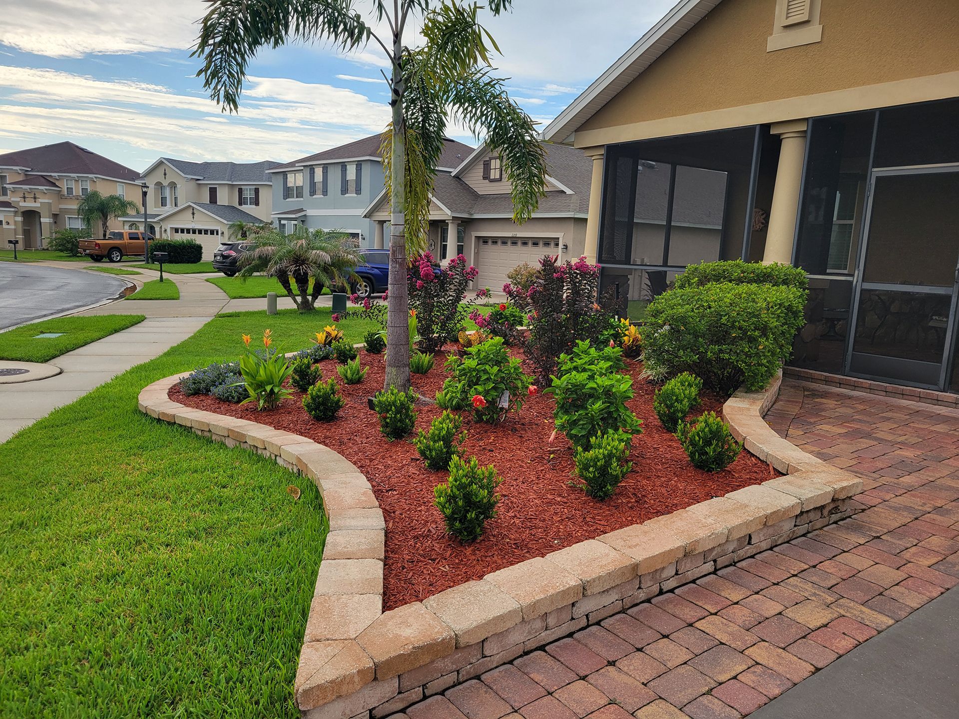 Front yard landscaping with red mulch, flowering shrubs, a small palm, and a brick walkway beside a house