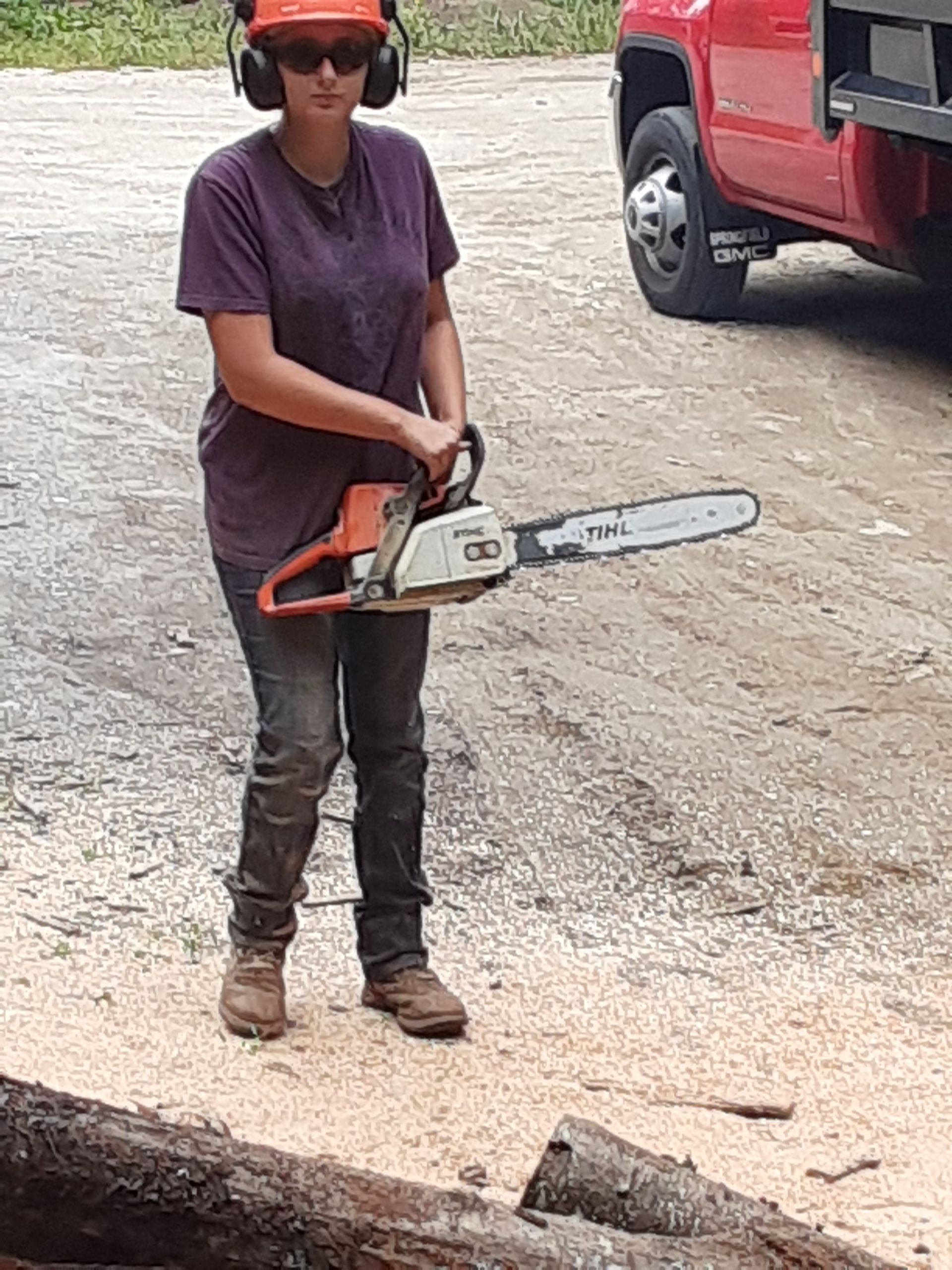 Woman wearing safety gear holding a chainsaw, standing near a red truck and logs.