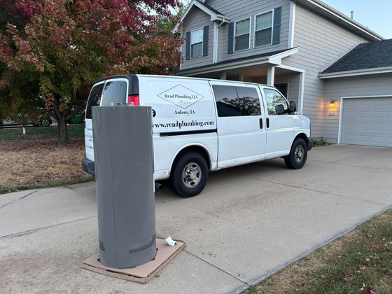 White van parked on driveway with gray cylindrical object on cardboard base in front of it, near a house.