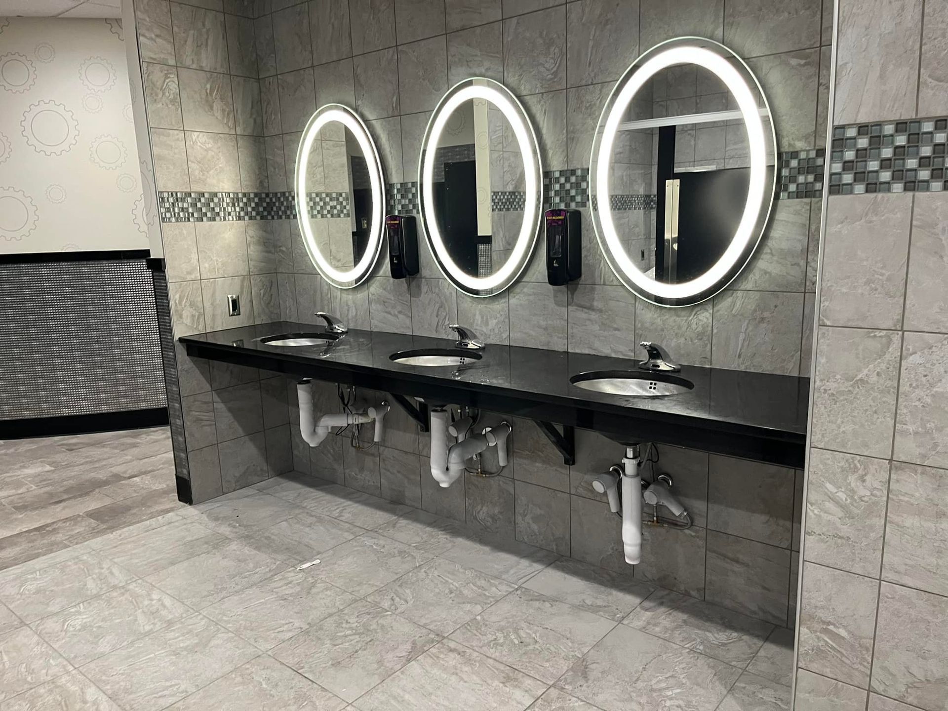 Three sinks with oval, lighted mirrors in a public restroom with tile walls and floor.
