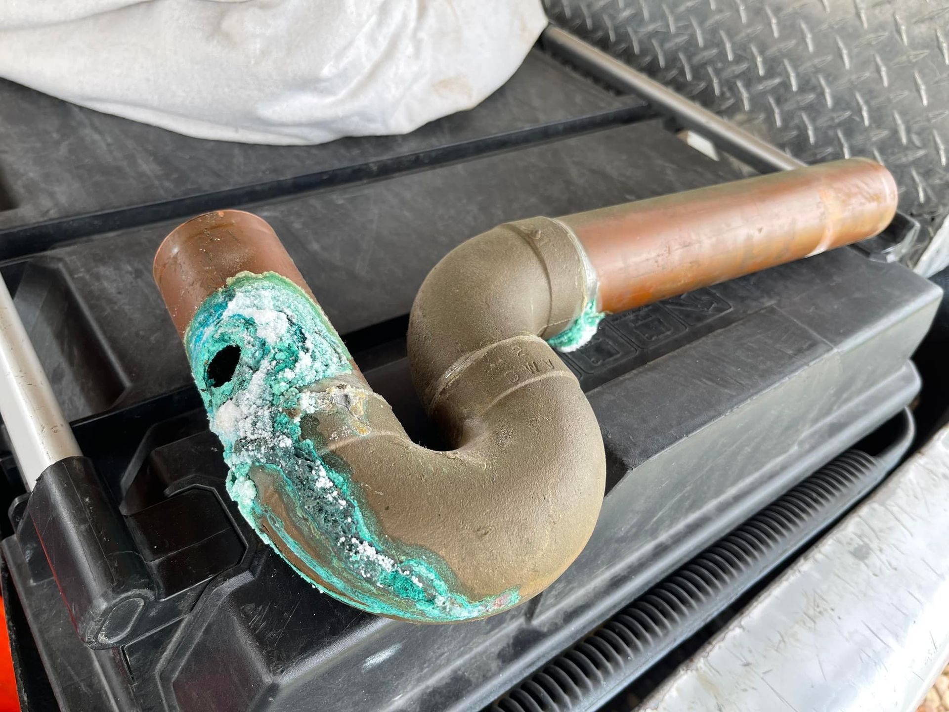 Copper pipe with heavy blue-green corrosion, lying on a black plastic container.