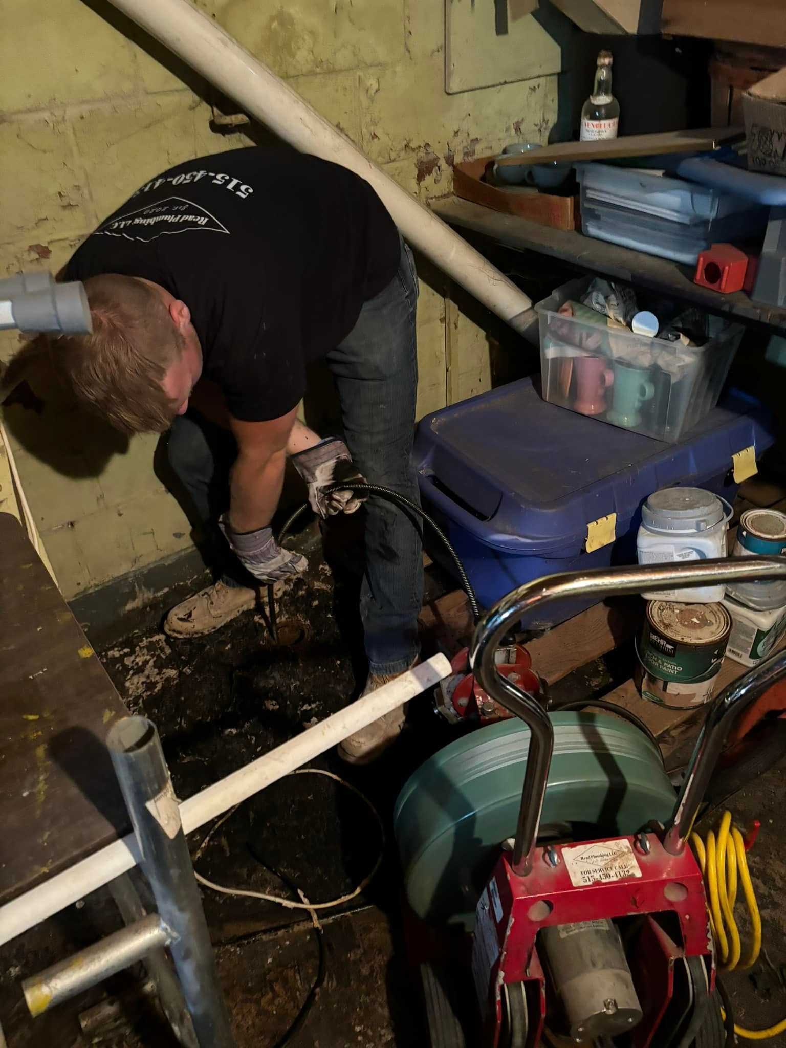 Man working on plumbing in a basement, using a machine. Dark setting with pipes and equipment visible.