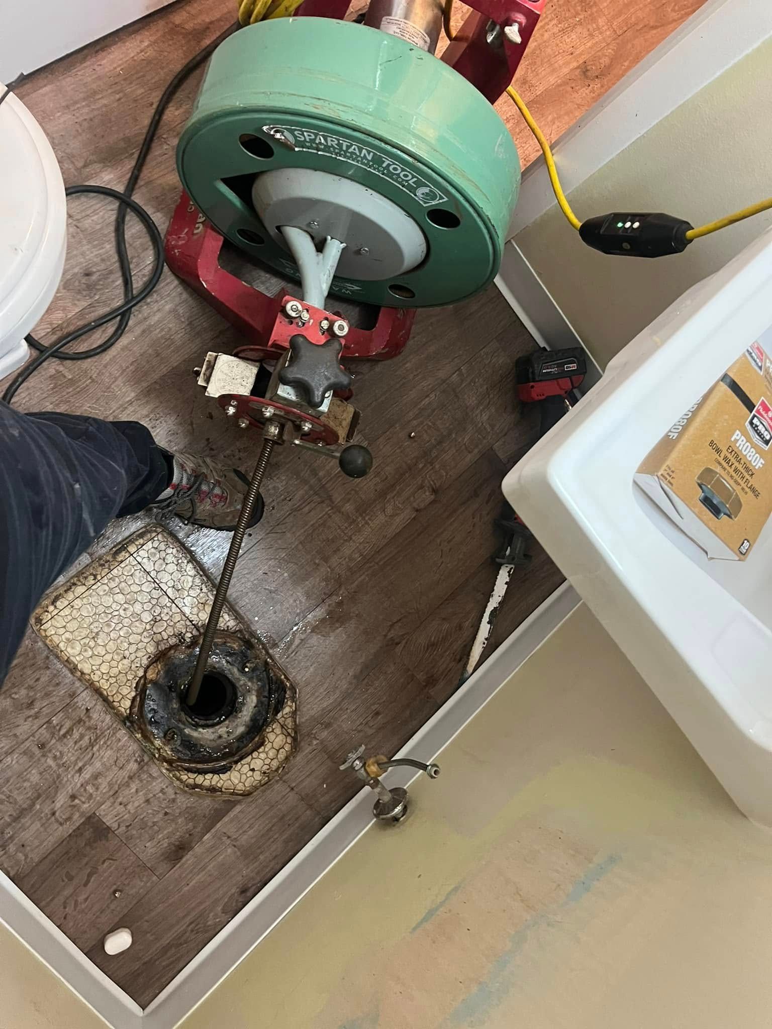 A drain cleaning machine next to a removed toilet, on a floor with wood-look tiles.