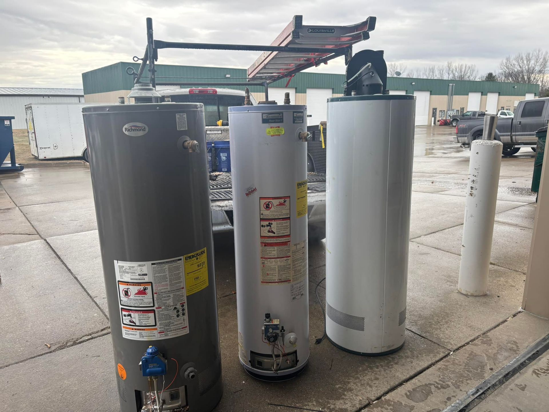 Three water heaters of different colors stand outdoors near a truck with a ladder rack.