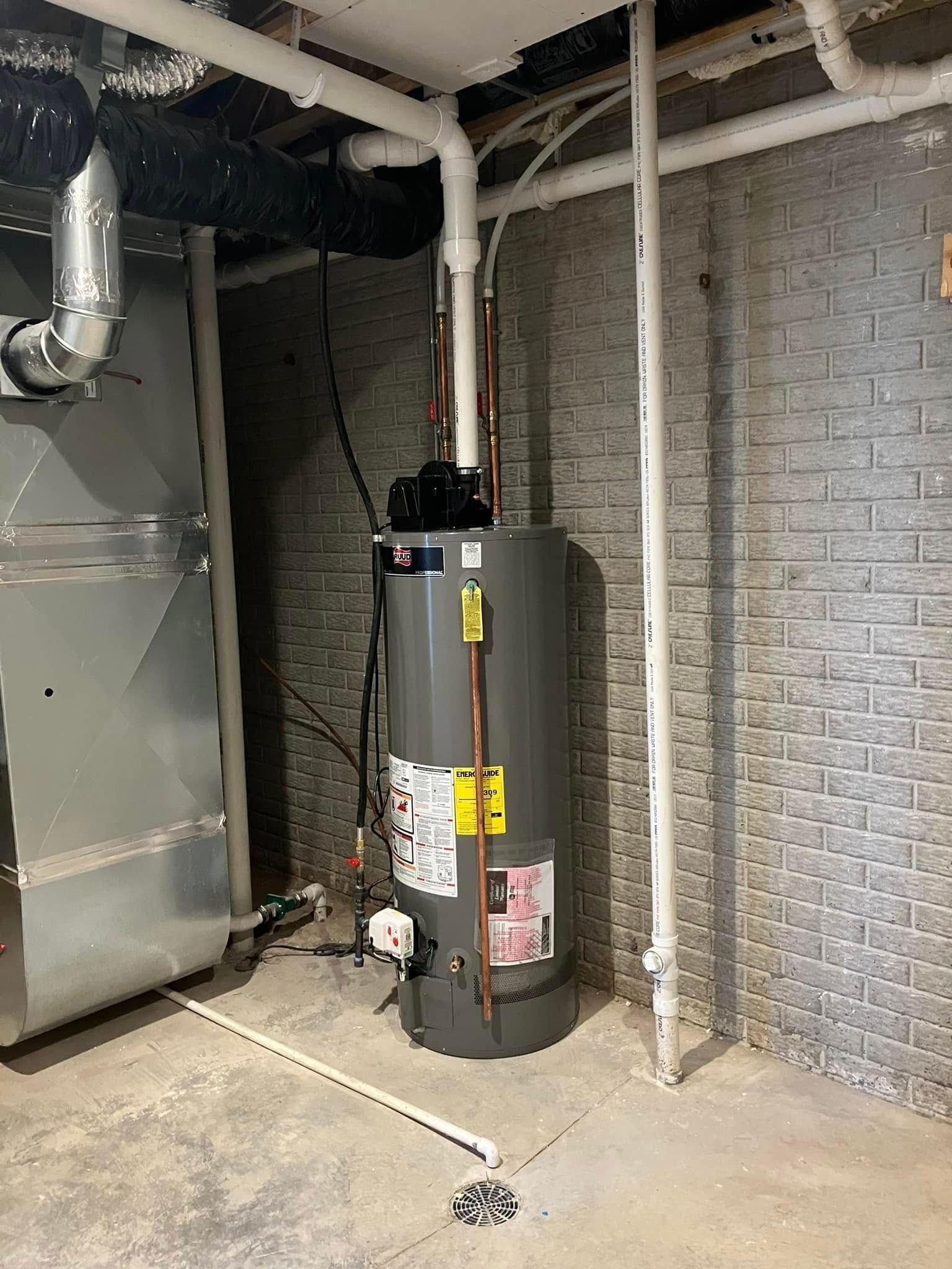 Water heater in a basement. It's gray with white pipes and a furnace to the left.