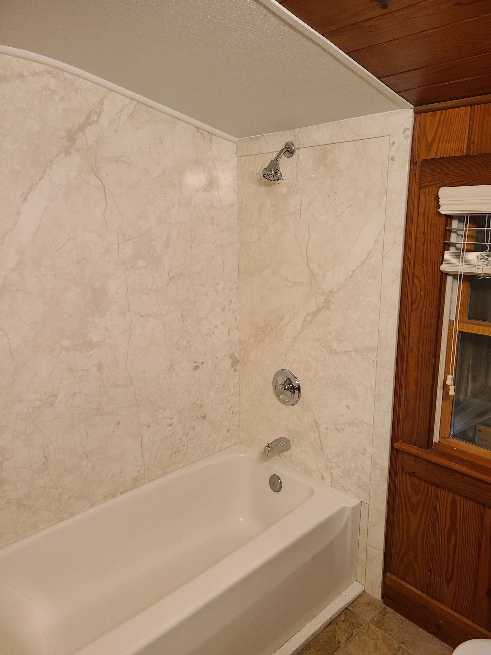 Bathtub with marble-like shower surround, showerhead, and faucet, next to a wood-paneled wall with a window.