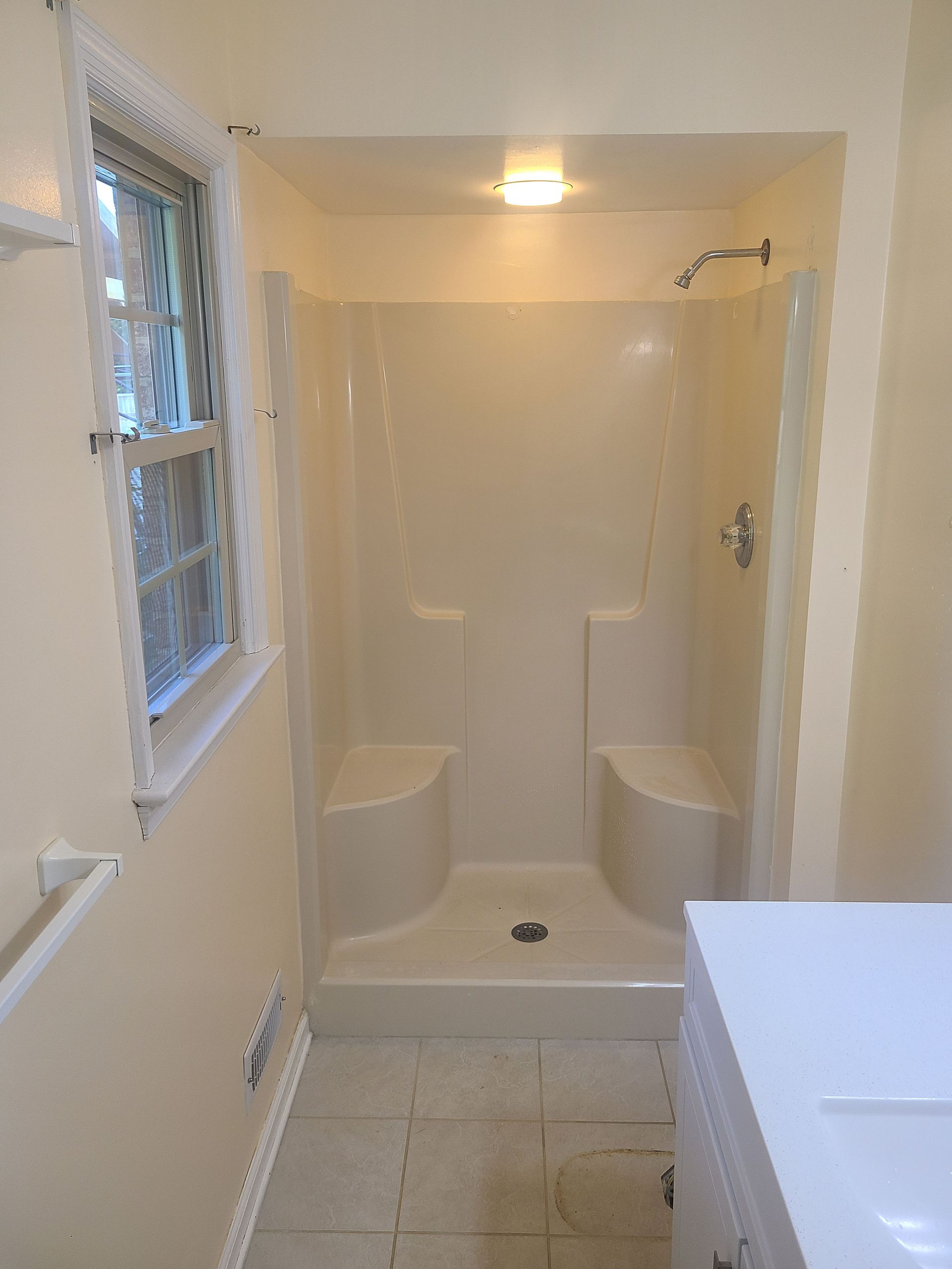 White bathroom with a shower, sink, window, and tiled floor.