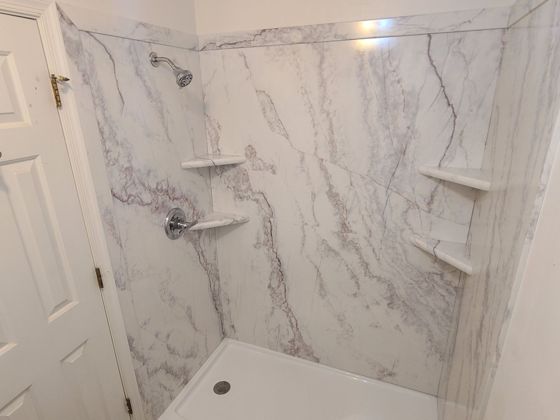 White marble-patterned shower with built-in shelves and a white door to the left.