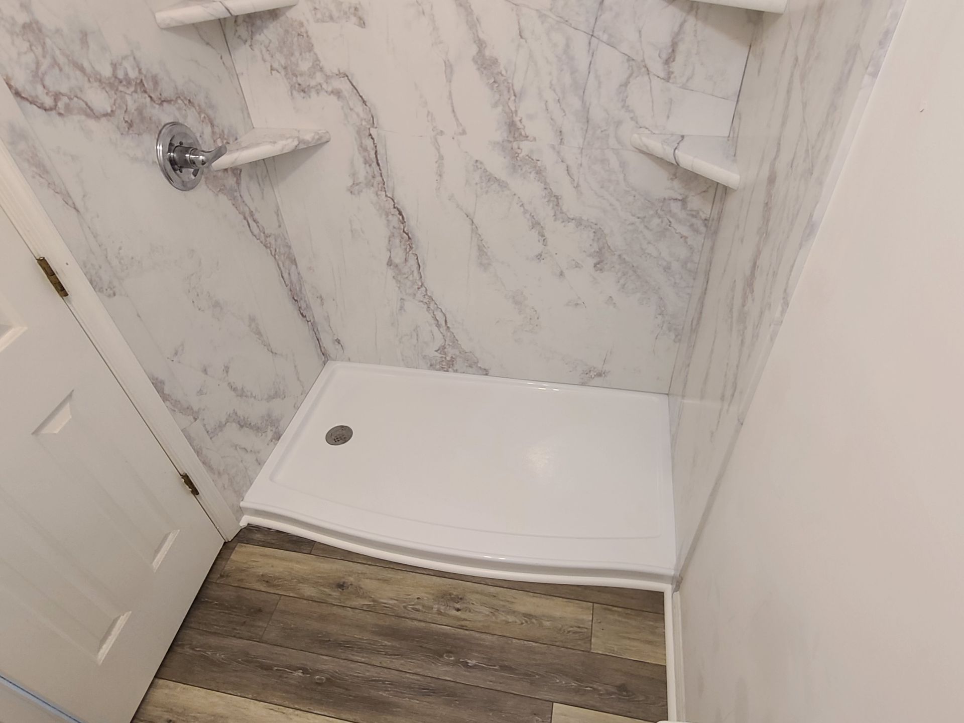 Shower stall with white marble-look walls, white shower base, chrome fixtures, and corner shelves.