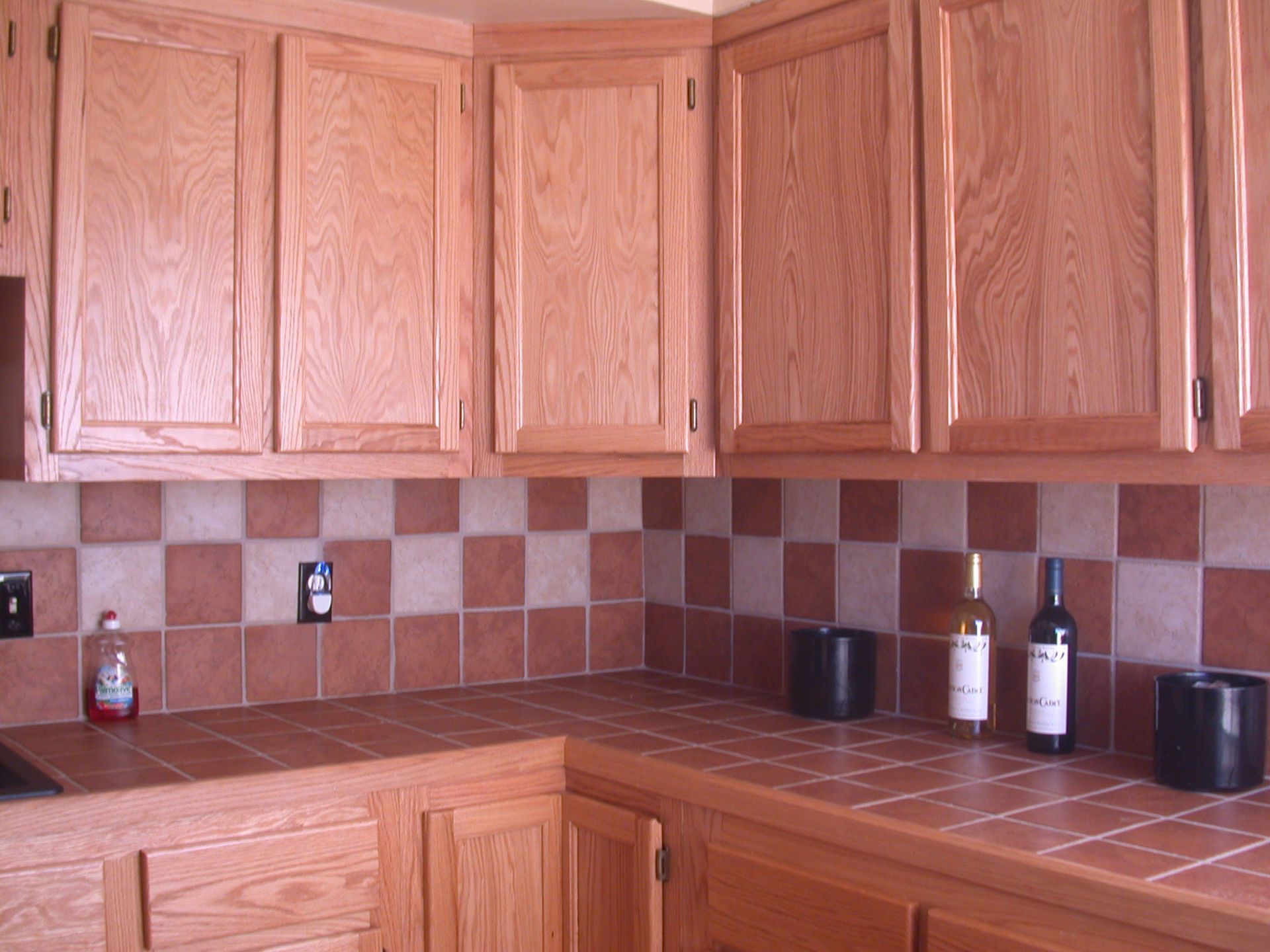 Oak kitchen cabinets with reddish-brown tiled backsplash and countertop. Bottles sit on the counter.