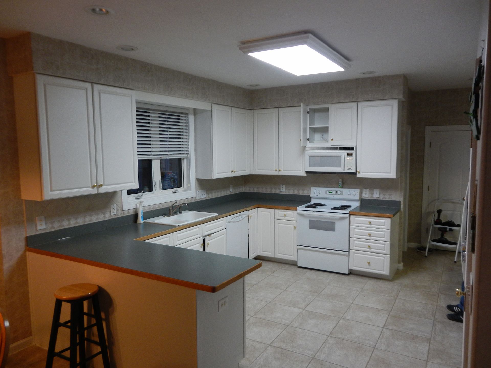 White kitchen with cabinets, appliances, and a countertop bar; a stool is in front.