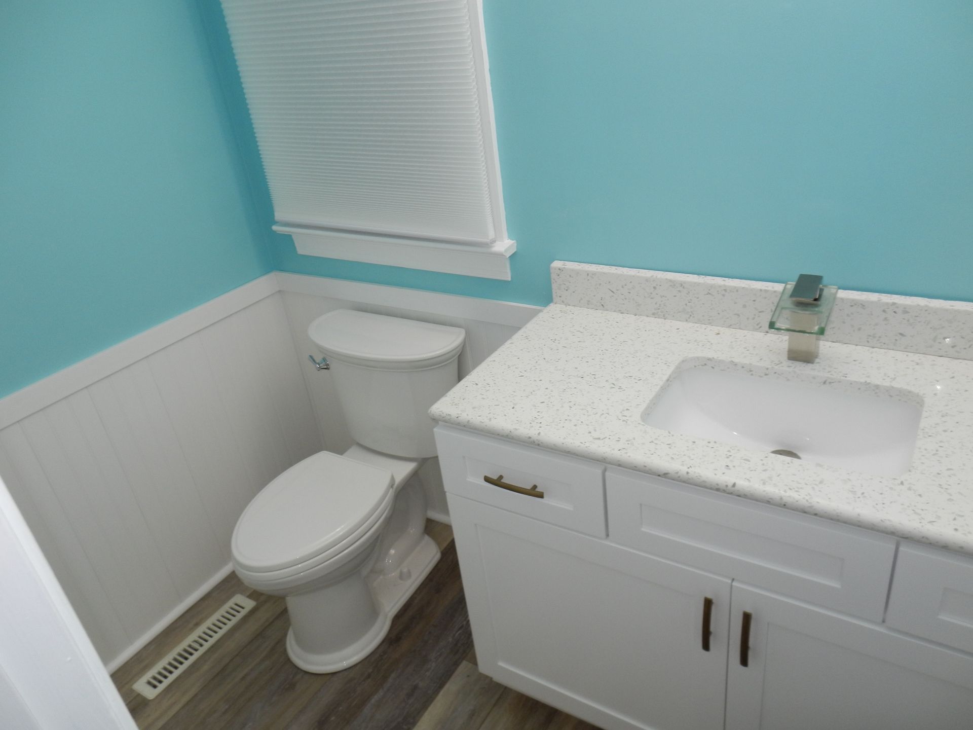 Bathroom with white vanity, toilet, and beadboard against blue walls, light-colored countertop, and blinds.