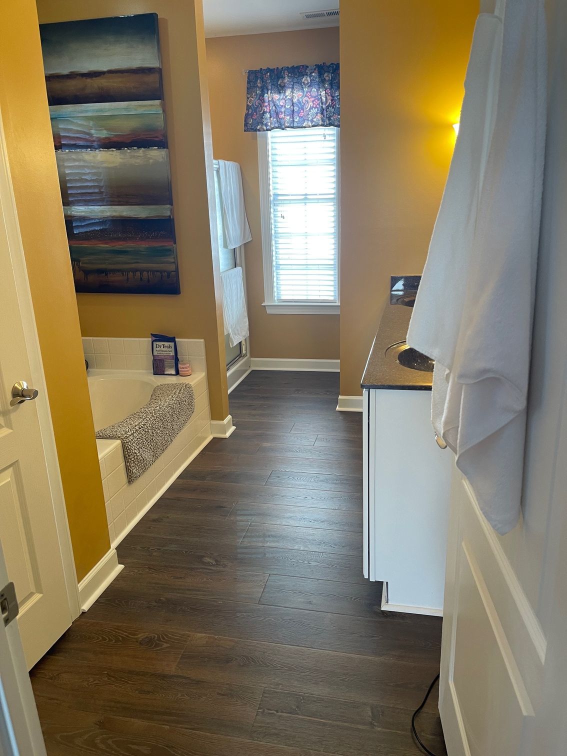 Bathroom with brown wood floor, white cabinets, and art on mustard walls. Bathtub and window are visible.