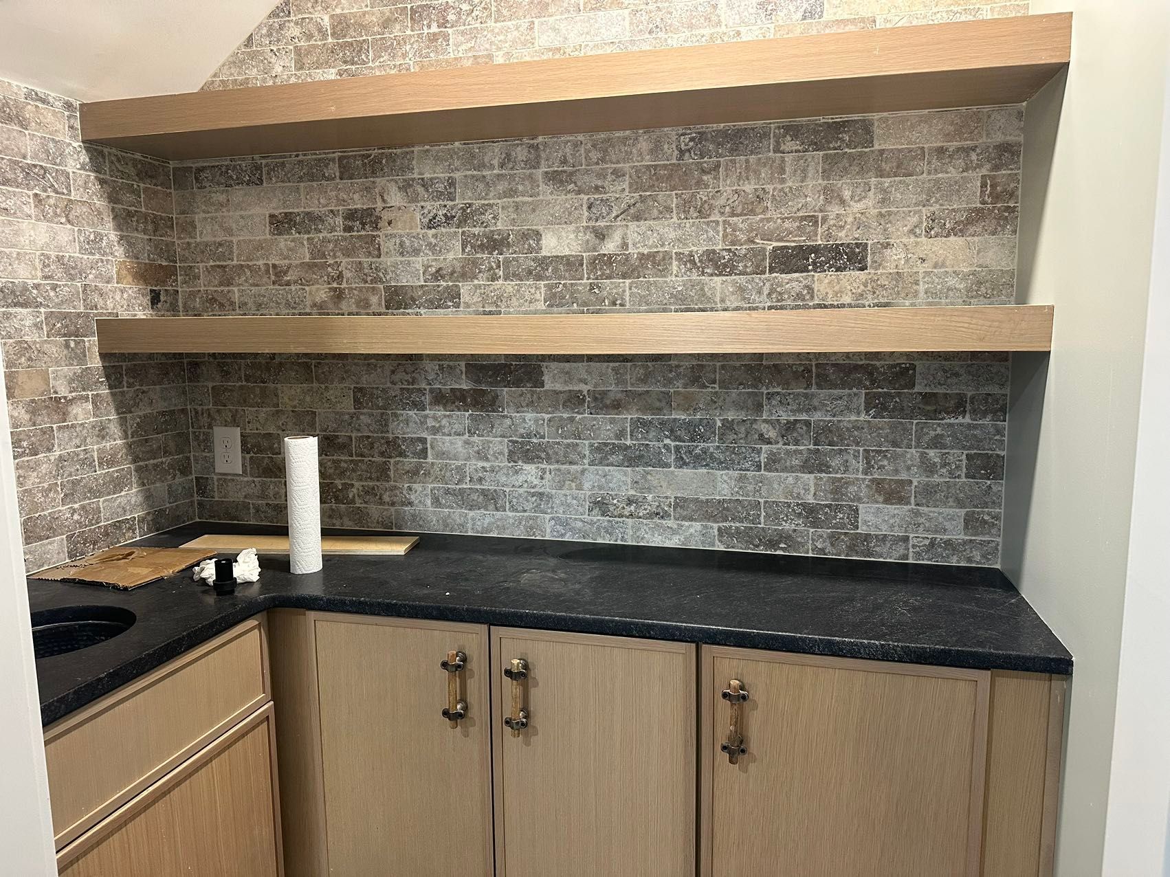 A kitchen with a sink, cabinets, shelves, and a brick wall.