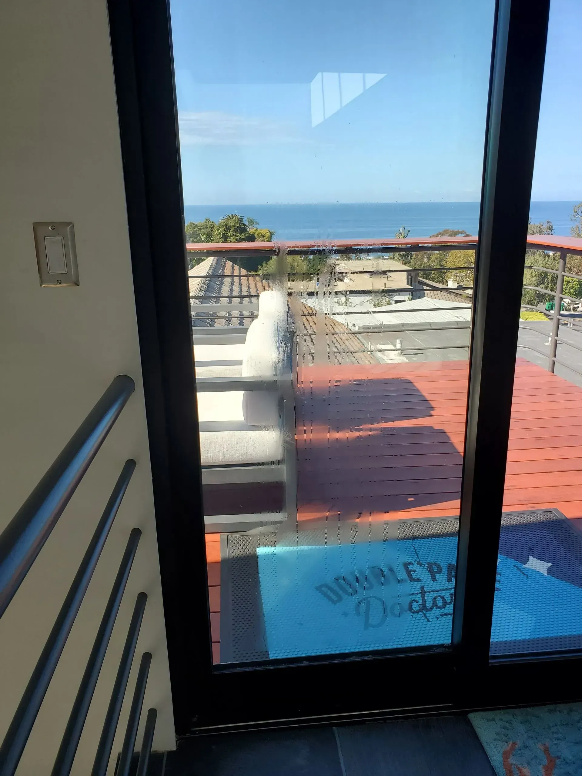 View through a sliding glass door to a rooftop deck with ocean view, blue mat, and seating.