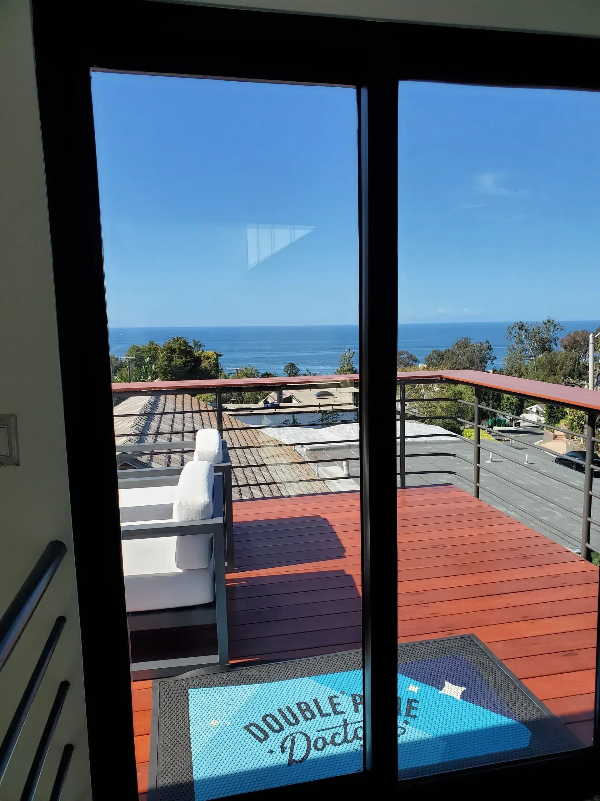 A balcony with ocean view seen through sliding glass doors. Red deck with white seating and a blue welcome mat.