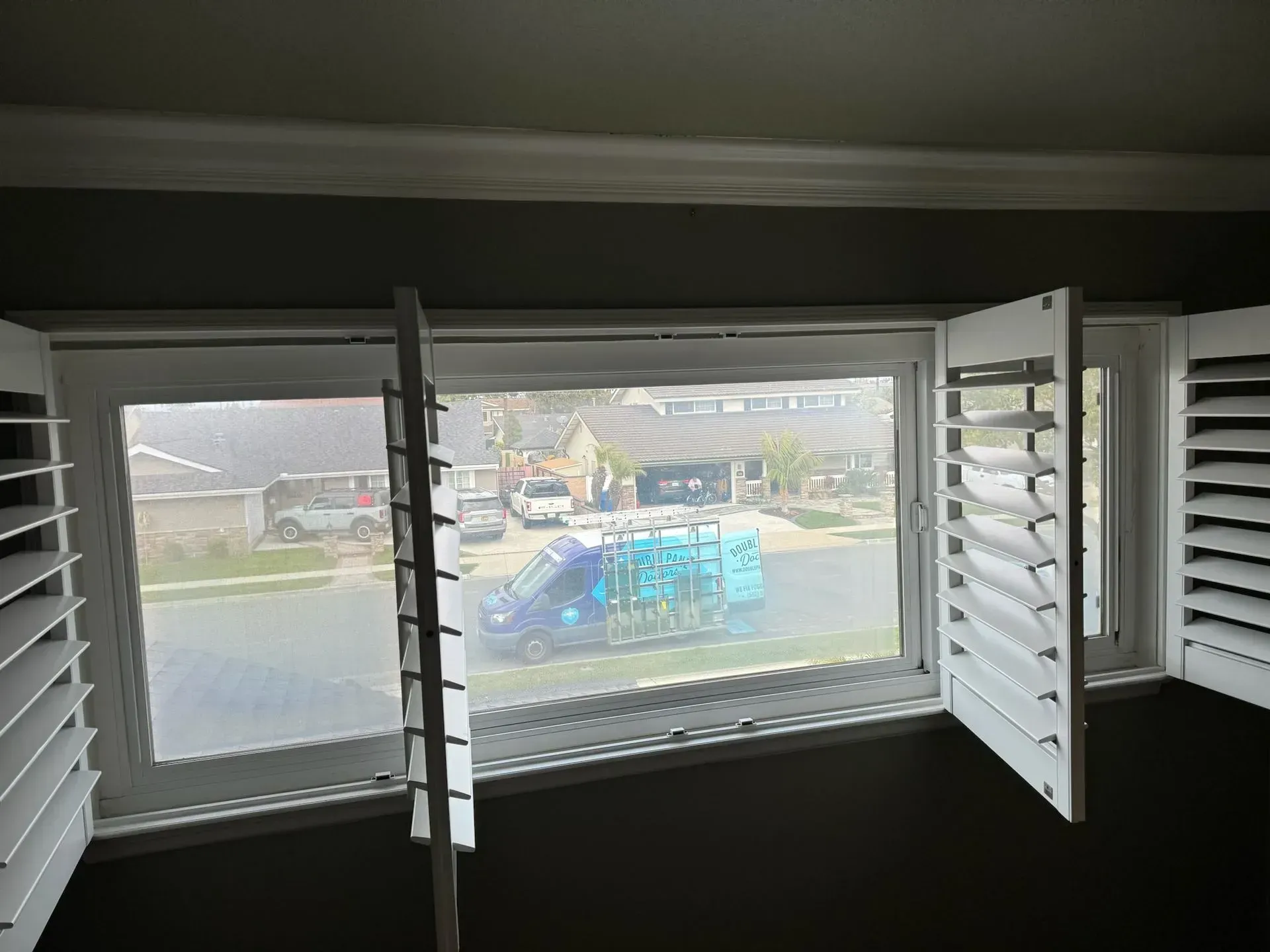 White window shutters are open, looking out onto a street scene with parked cars and a van.