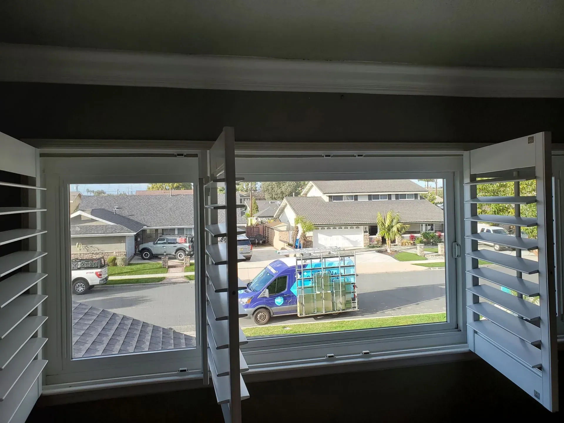 Open white shutters in a home window, overlooking a street with houses and a blue van.