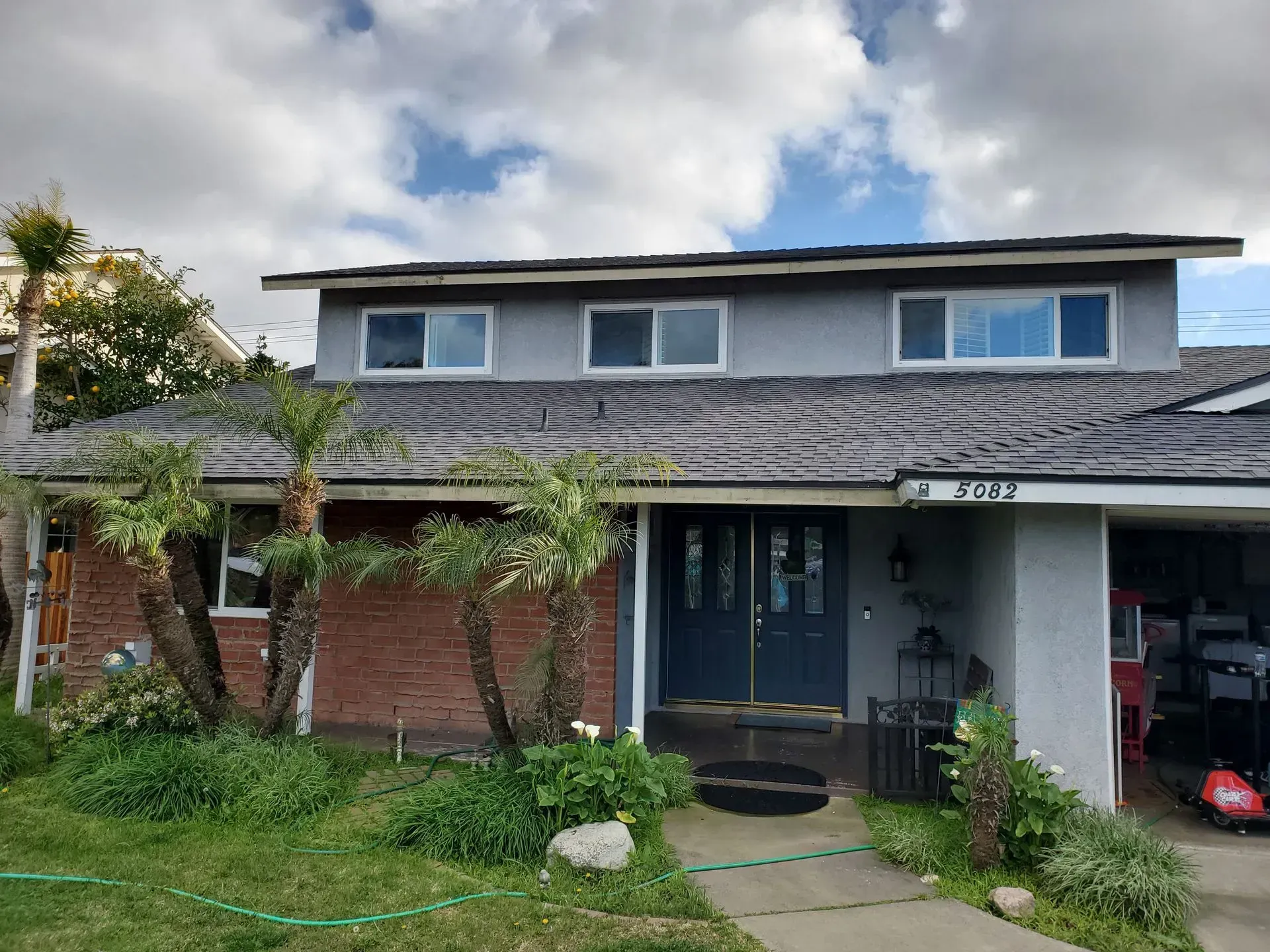 Two-story house with gray stucco and red brick, blue door, palm trees, green grass, and cloudy sky.