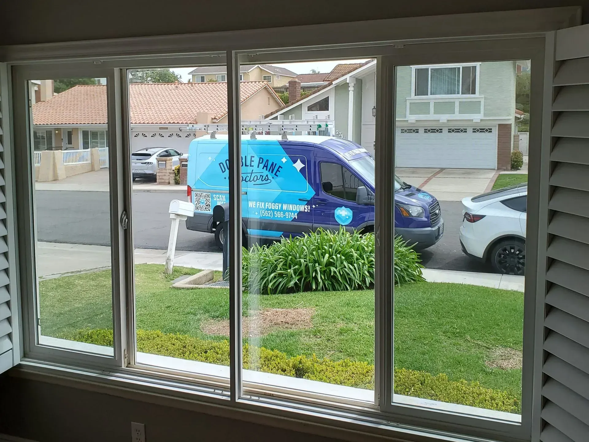 View through a window of a blue van parked on a residential street with a green lawn and houses.