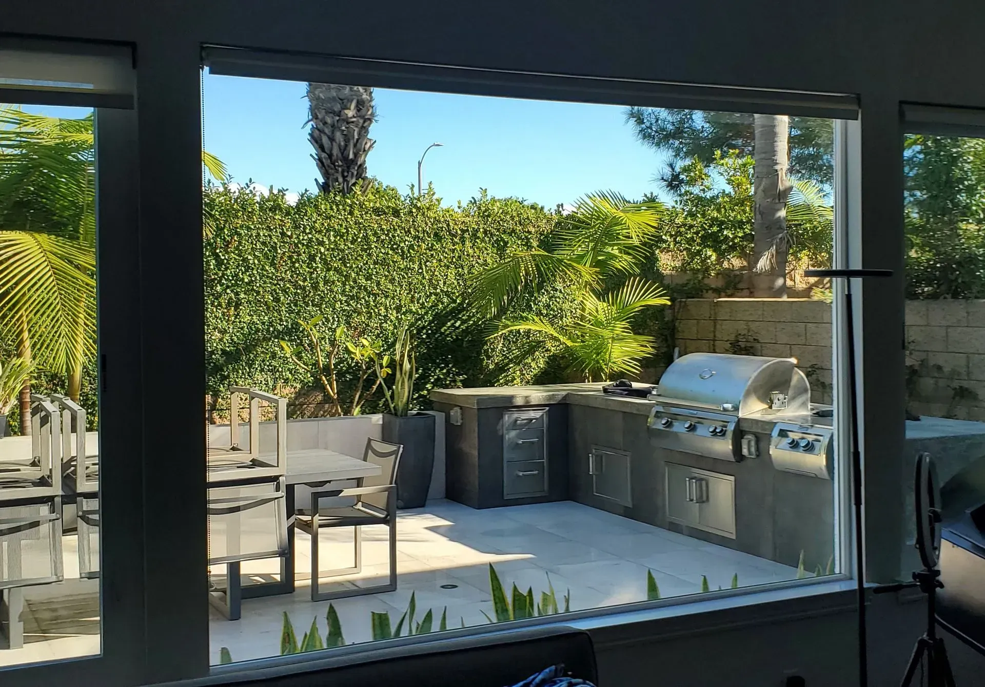 Outdoor patio with grill, table, and lush greenery, viewed through a window.
