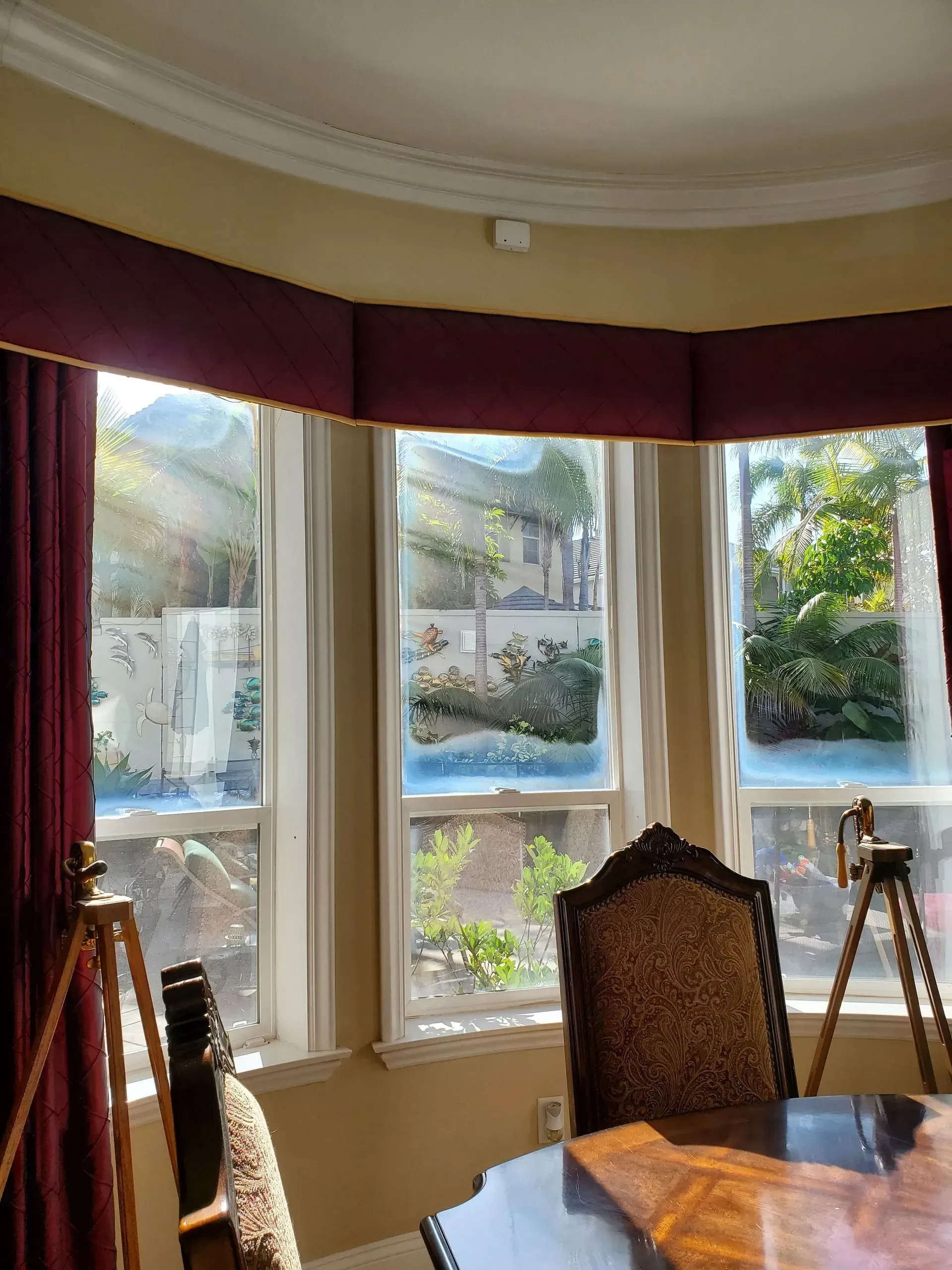 Bay window with red valance and curtains, overlooking a garden. A table and chairs are in the foreground.