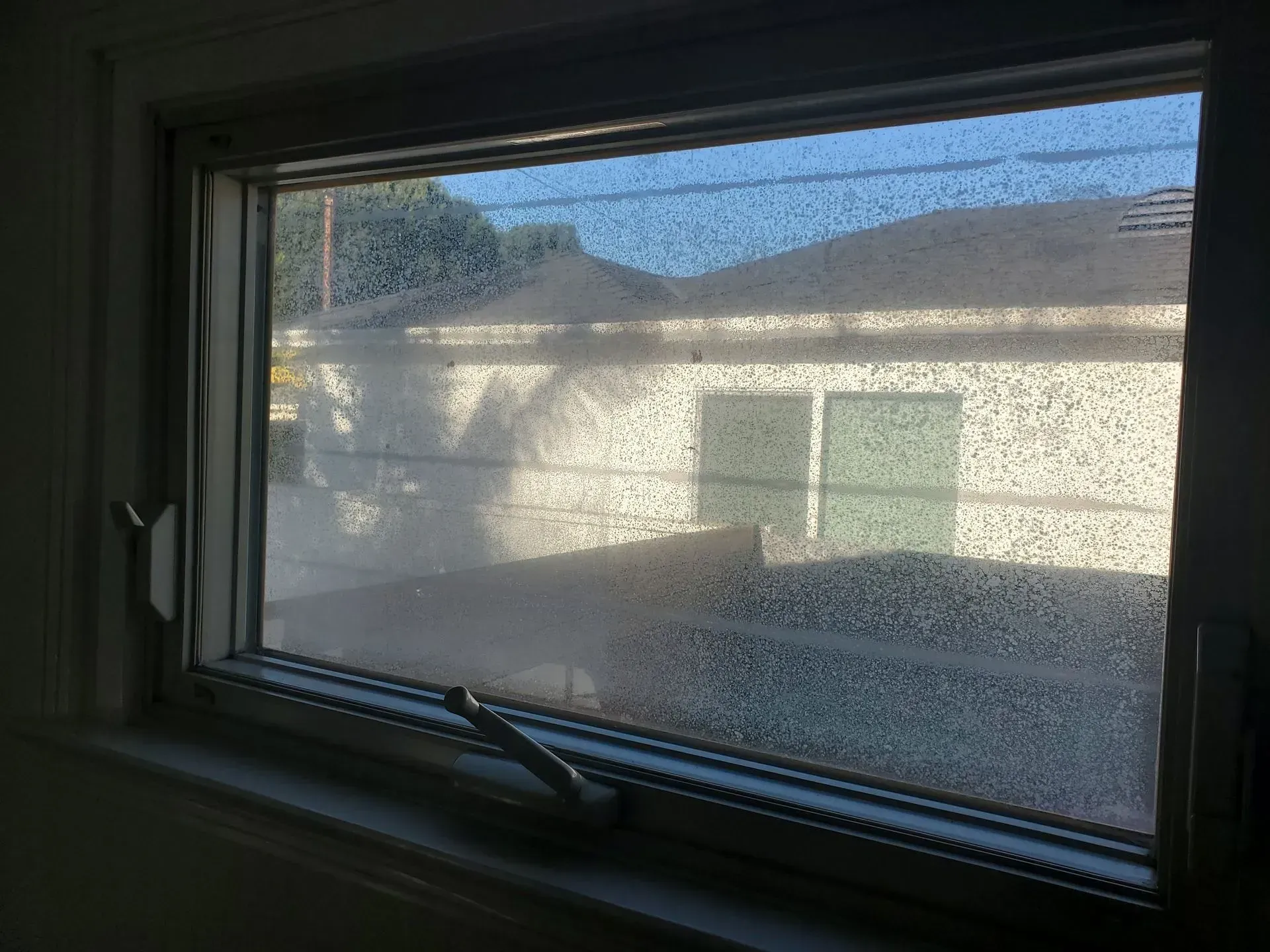 Window with condensation, looking out to a backyard with a house and blue sky.