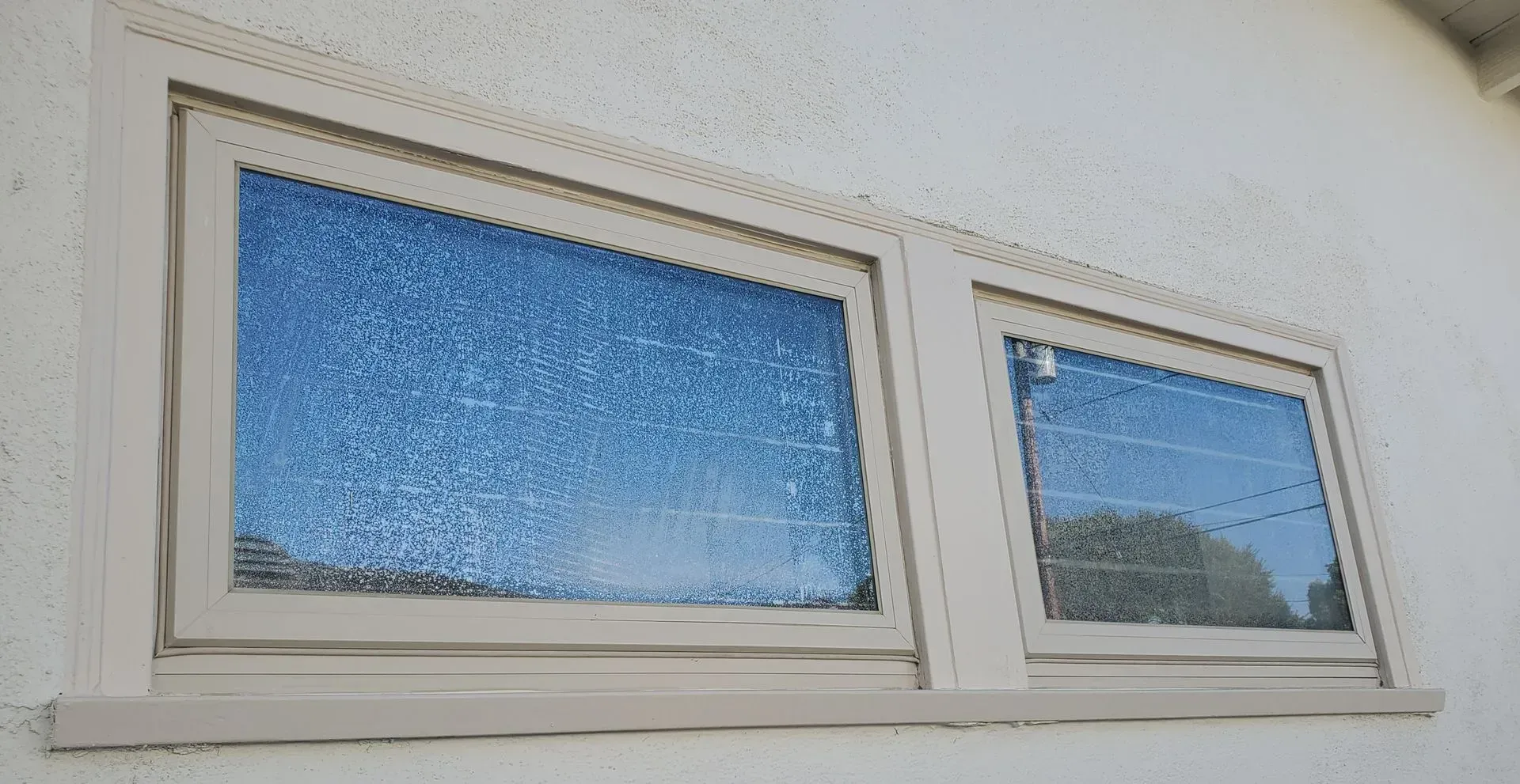 Two beige-framed windows on a white building, reflecting a blue sky and trees.