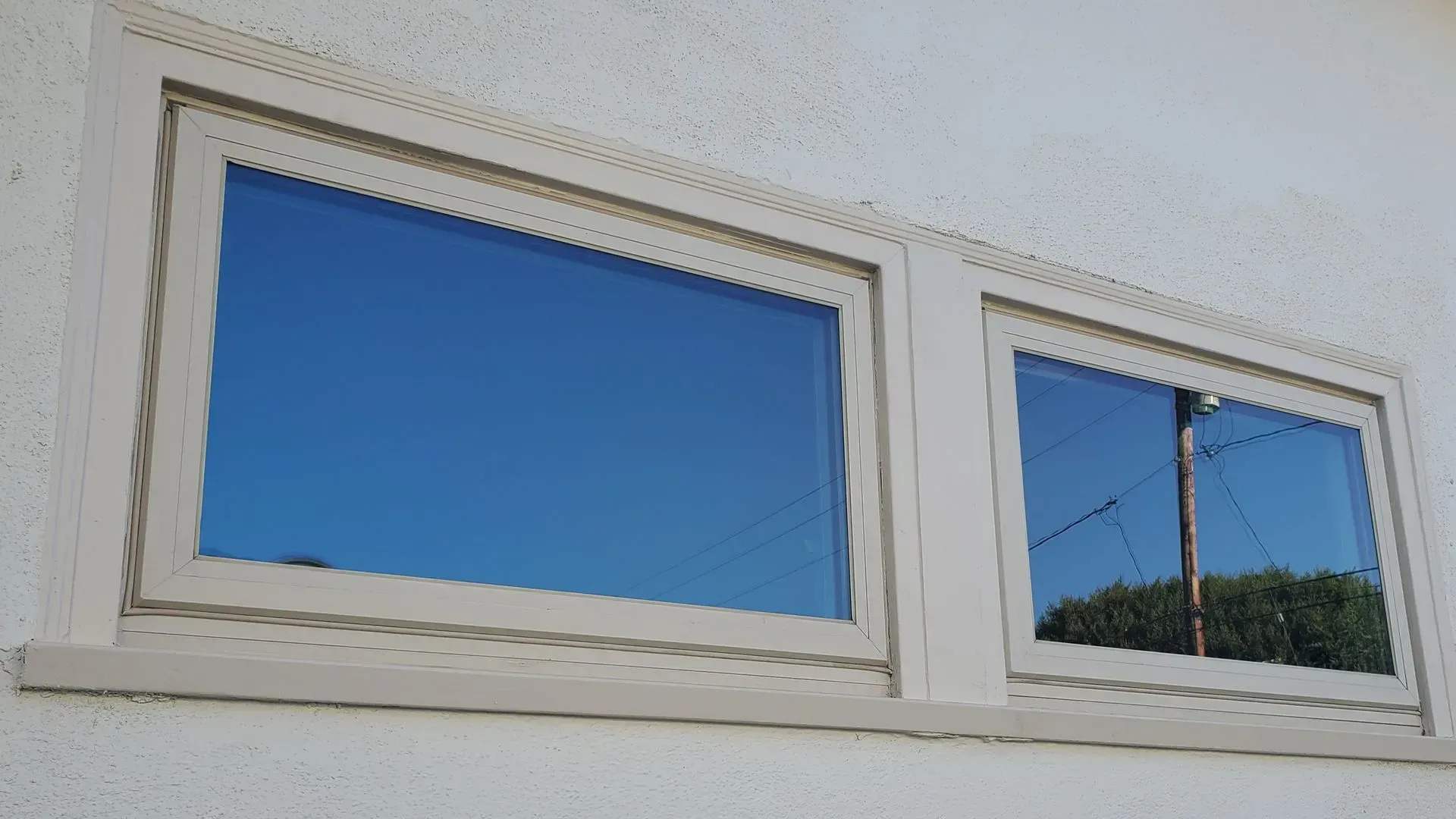Two beige-framed windows reflecting a bright blue sky, set in a white stucco wall.