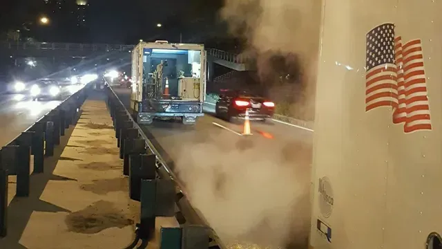 Steam billowing near a truck and a car on a road, night scene. American flag on a vehicle.