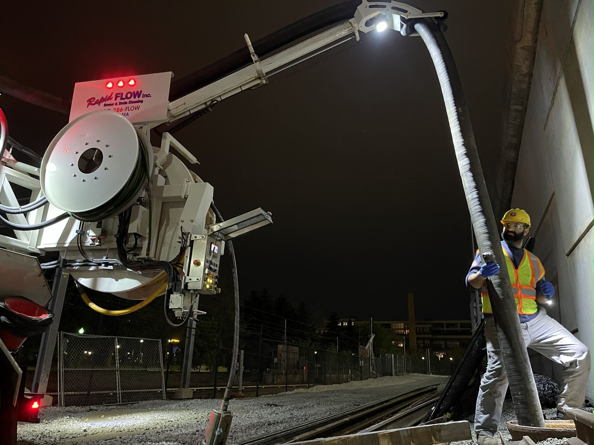 Worker operating a machine near train tracks at night. The machine has a long arm with a light, a reel, and a worker holds a hose.