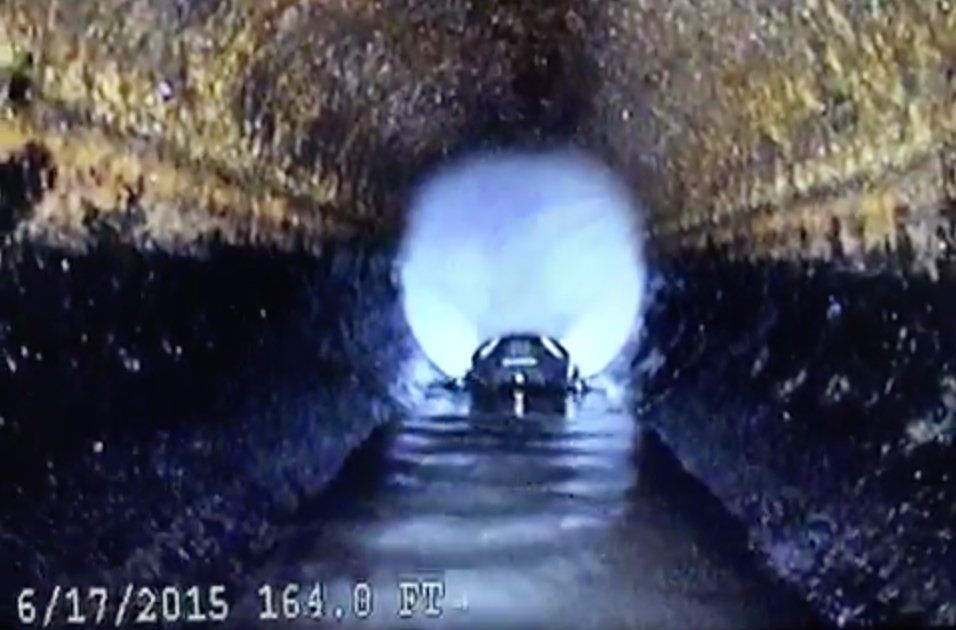 Camera view inside a sewer pipe, water flowing, light at the end, date stamp.
