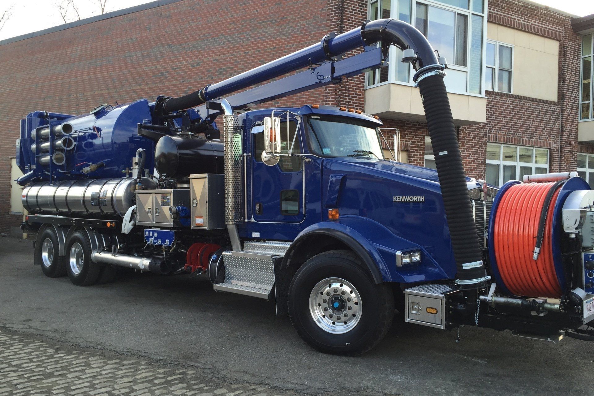Blue vacuum truck parked on brick street. Dark hose and reel on the right. Large tank and boom arm.