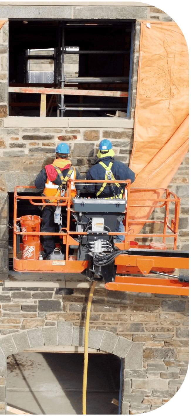 Two workers in orange lift at a stone wall opening, wearing hard hats and safety gear