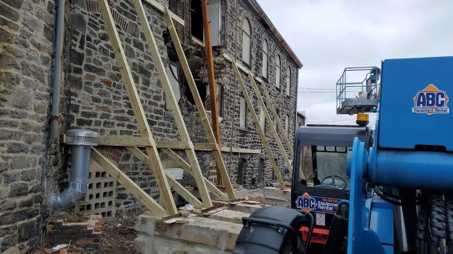 Exterior of a stone building undergoing repair. Wooden bracing and a lift are visible. Cloudy sky.