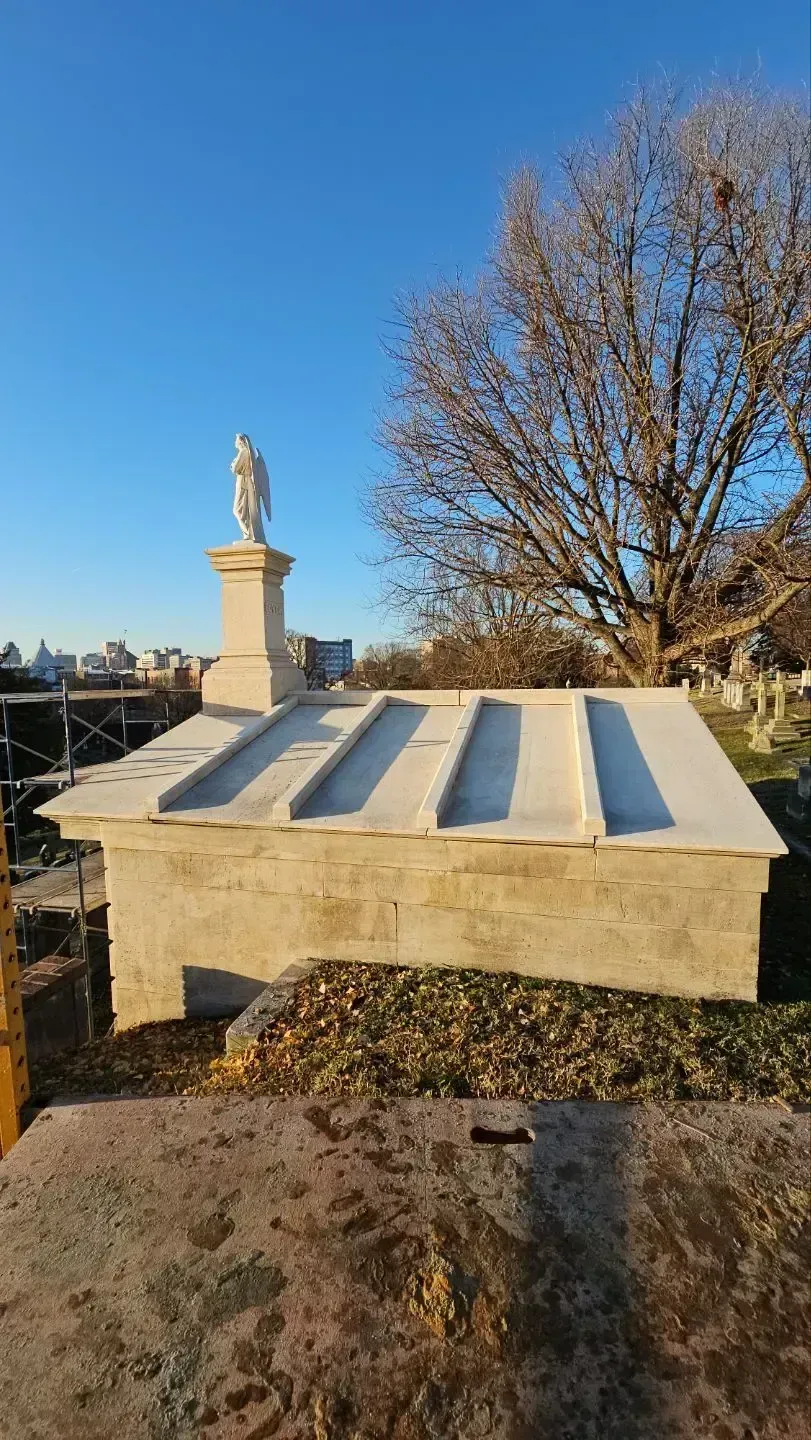 Stone mausoleum with statue, blue sky, and leafless tree.