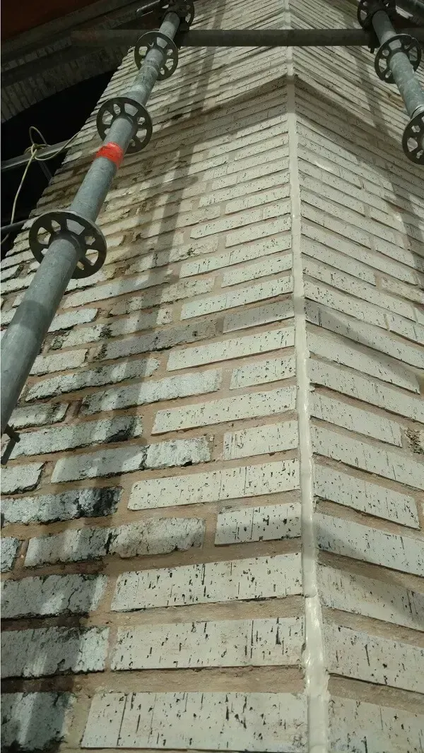 Brick wall corner with scaffolding; light-colored bricks, mortar, and gray metal support.