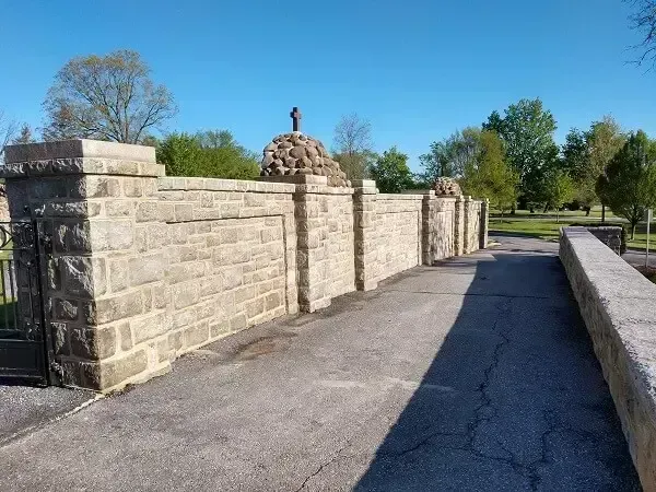 Stone wall with cross-topped structure and paved path in a park-like setting under a blue sky.