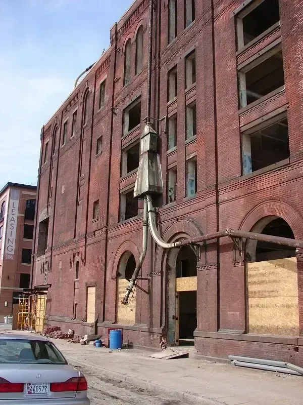 Red brick building exterior with boarded-up windows, pipes, and a car parked in front.