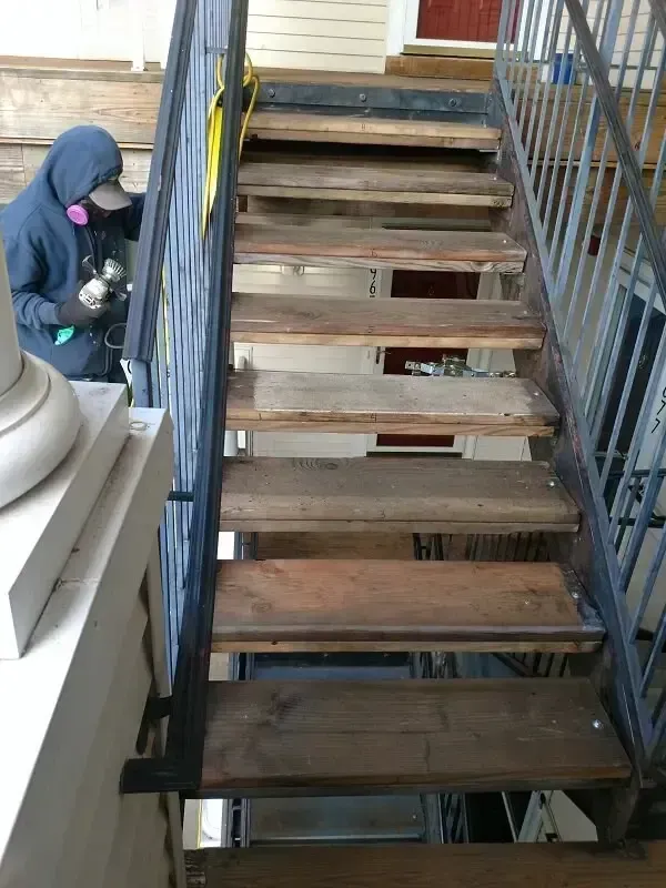 Person spraying brown stain on wooden exterior staircase. Metal railing and safety mask visible.