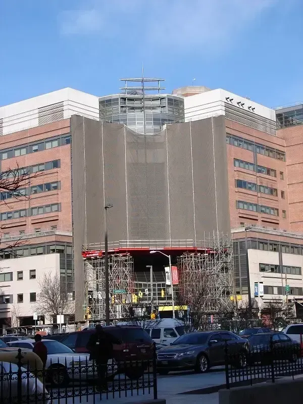 Hospital building with construction scaffolding and netting. Cars and people in the foreground.