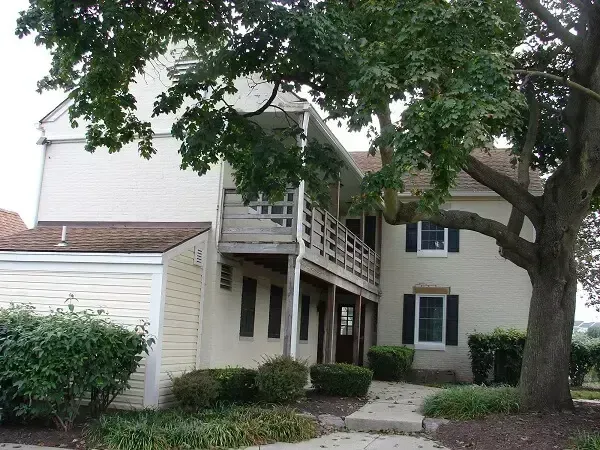 Two-story beige building with brown roof and wooden balcony, obscured by a large tree.